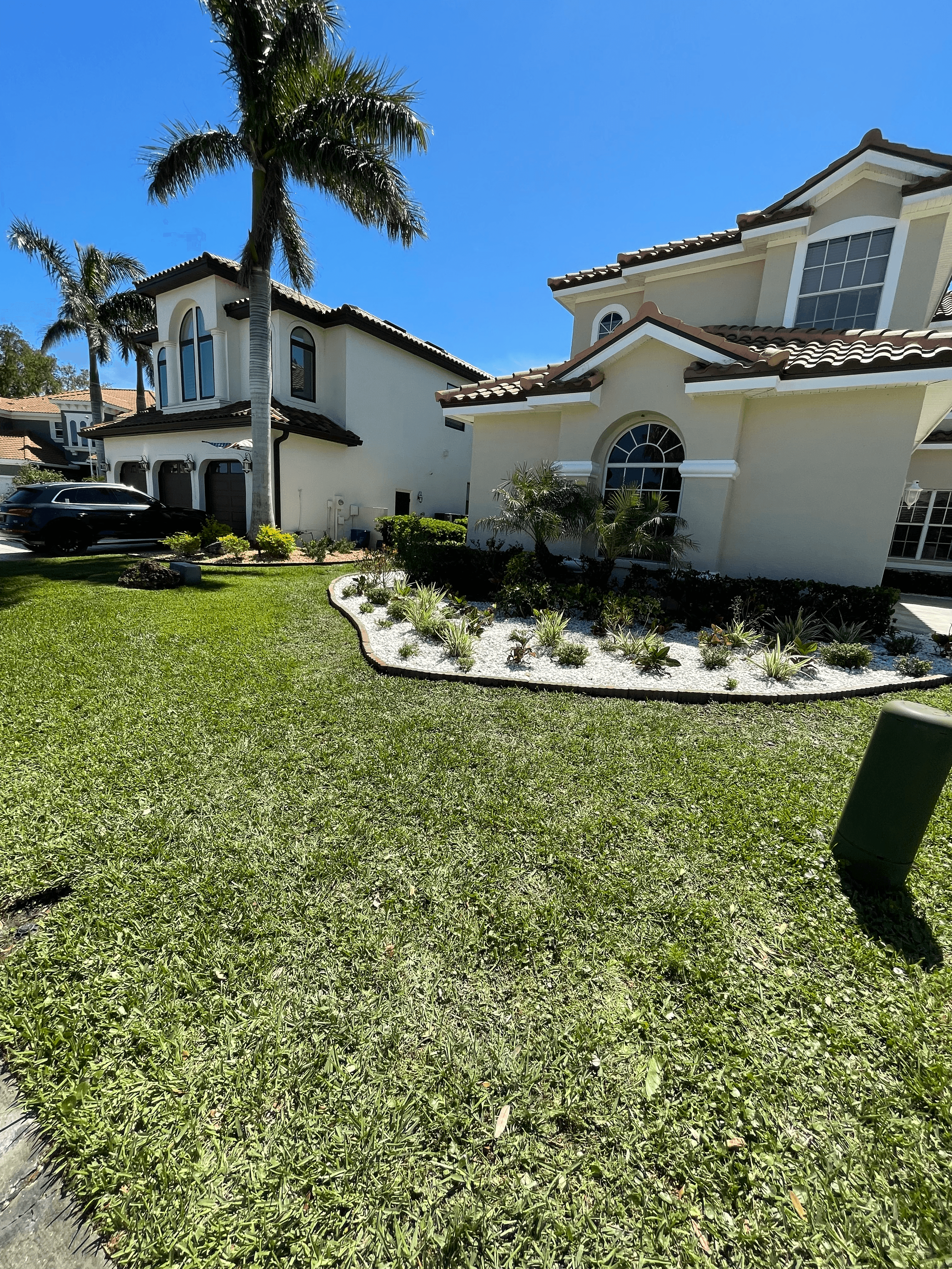 Beige two-story house with tile roof, palm tree, and white rock garden on green lawn.